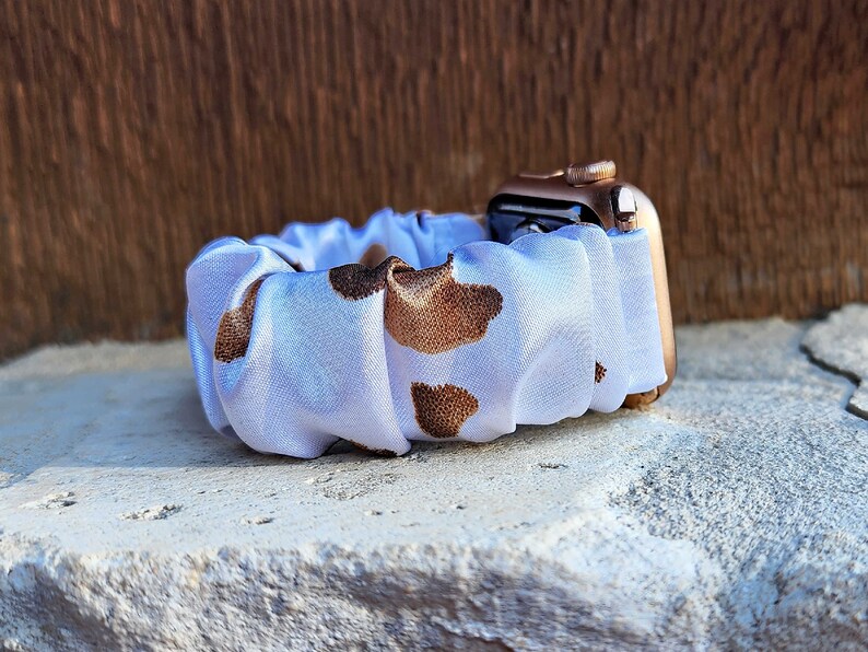 Op de afbeelding: Een horlogeband in scrunchie-stijl met een witte stoffen basis en een bruin koeienvlekkenpatroon. De band is bevestigd aan een ros&eacute;gouden smartwatch-behuizing en heeft een geplooid, elastisch ontwerp dat om de pols past.