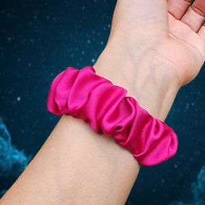 May include: A bright pink satin scrunchie, worn on a person's wrist.