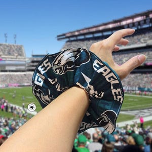 Puede incluir: Una scrunchie verde azulado y blanca con el logotipo y el texto de los Eagles de Filadelfia. La scrunchie se lleva en la mu&ntilde;eca, con un estadio de f&uacute;tbol al fondo.