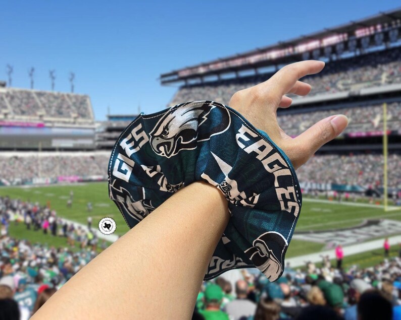 Puede incluir: Una scrunchie verde azulado y blanca con el logotipo y el texto de los Eagles de Filadelfia. La scrunchie se lleva en la mu&ntilde;eca, con un estadio de f&uacute;tbol al fondo.