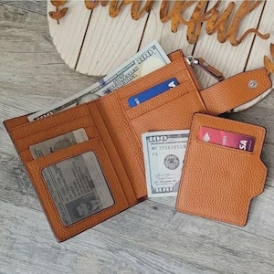 May include: Open, tan leather wallet with multiple card slots, a clear ID window, and a separate card holder. The wallet contains cash and credit cards. The wallet is open, displaying its contents.