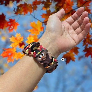 Op de afbeelding: Een bruin en zwart scrunchie met een cartoon uil ontwerp, met een zwarte hoed en een rood en geel patroon. De scrunchie zit om een pols, tegen een achtergrond van herfstbladeren.