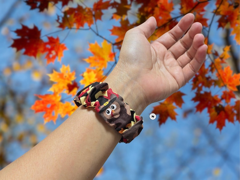 Op de afbeelding: Een bruin en zwart scrunchie met een cartoon uil ontwerp, met een zwarte hoed en een rood en geel patroon. De scrunchie zit om een pols, tegen een achtergrond van herfstbladeren.