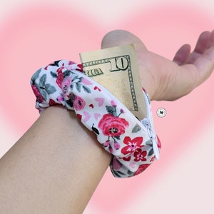 Peut inclure: Un portefeuille de poignet floral avec un fond blanc, avec des roses rouges et roses et de petits c&oelig;urs. Le portefeuille est ouvert, r&eacute;v&eacute;lant un billet de dix dollars. Le portefeuille de poignet est con&ccedil;u pour &ecirc;tre port&eacute; autour du poignet.