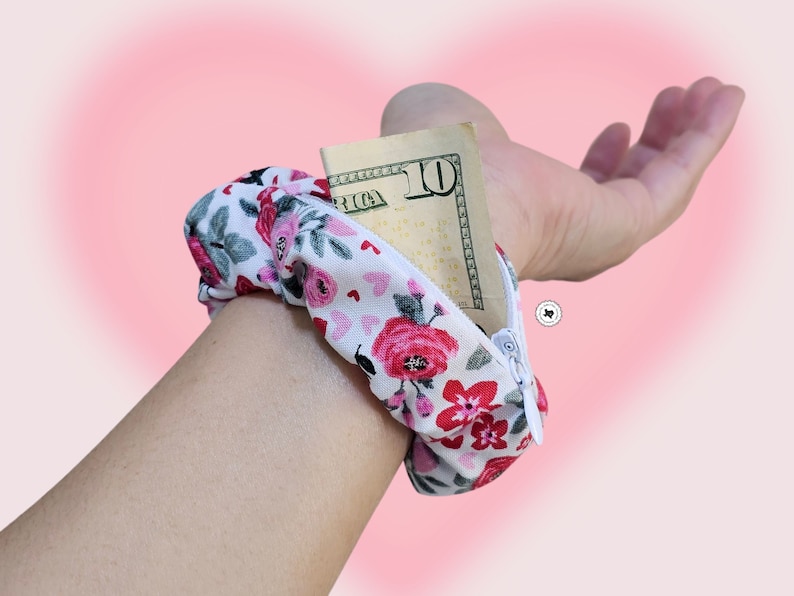 Peut inclure: Un portefeuille de poignet floral avec un fond blanc, avec des roses rouges et roses et de petits c&oelig;urs. Le portefeuille est ouvert, r&eacute;v&eacute;lant un billet de dix dollars. Le portefeuille de poignet est con&ccedil;u pour &ecirc;tre port&eacute; autour du poignet.