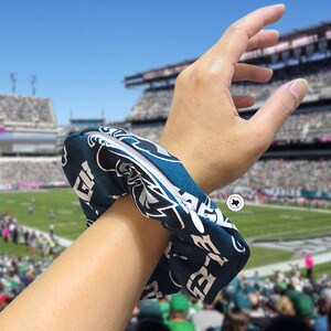 Puede incluir: Una scrunchie azul verdoso oscuro y blanco con el logotipo y el texto de los Philadelphia Eagles. La scrunchie est&aacute; alrededor de una mu&ntilde;eca, con un estadio de f&uacute;tbol americano al fondo.