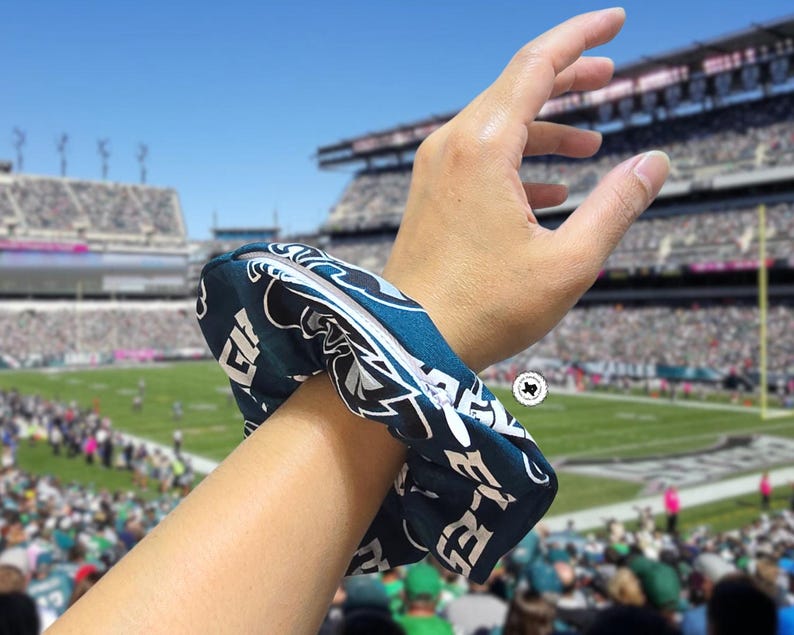 Puede incluir: Una scrunchie azul verdoso oscuro y blanco con el logotipo y el texto de los Philadelphia Eagles. La scrunchie est&aacute; alrededor de una mu&ntilde;eca, con un estadio de f&uacute;tbol americano al fondo.