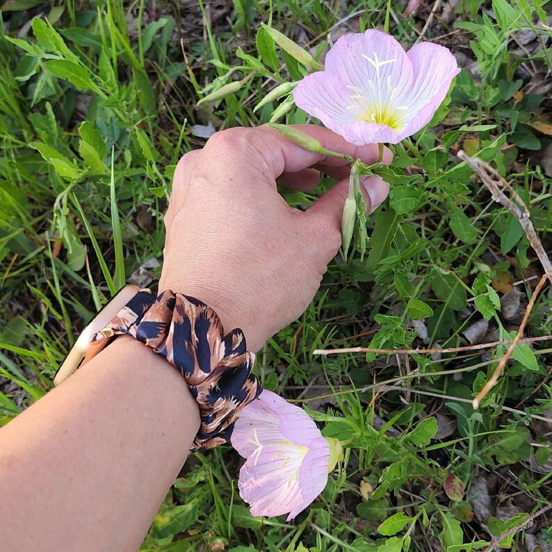 Op de afbeelding: Een horlogeband met een bruin en zwart luipaardpatroon en een geplooid stoffen ontwerp in scrunchie-stijl. De band is bevestigd aan een metalen horlogekast en wordt om de pols gedragen bij twee lichtroze bloemen met gele harten.