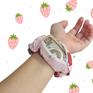 Peut inclure: Un chouchou rose avec un motif de fraises, dot&eacute; d'une poche zipp&eacute;e. La poche contient de l'argent et un petit objet rond. Le chouchou est port&eacute; au poignet sur un fond blanc avec des illustrations de fraises.