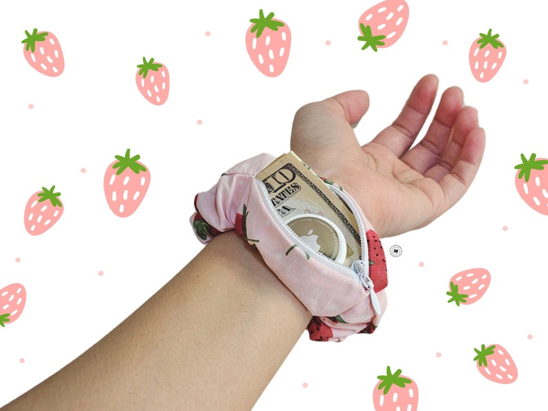 Peut inclure: Un chouchou rose avec un motif de fraises, dot&eacute; d'une poche zipp&eacute;e. La poche contient de l'argent et un petit objet rond. Le chouchou est port&eacute; au poignet sur un fond blanc avec des illustrations de fraises.