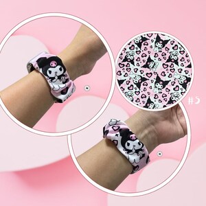 Op de afbeelding: Roze en witte scrunchie met een zwart cartoonkarakterontwerp. De scrunchie wordt getoond op een pols, met een close-up van het patroon. Het patroon bevat harten en het cartoonkarakter.