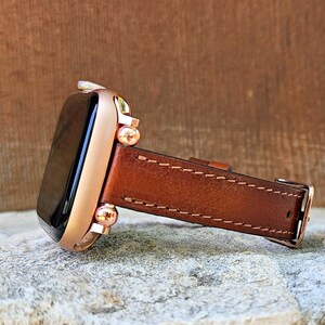 May include: A rose gold smartwatch with a brown leather band. The watch face is black, and the band is stitched with a lighter brown thread. The watch is displayed on a textured, light gray surface.