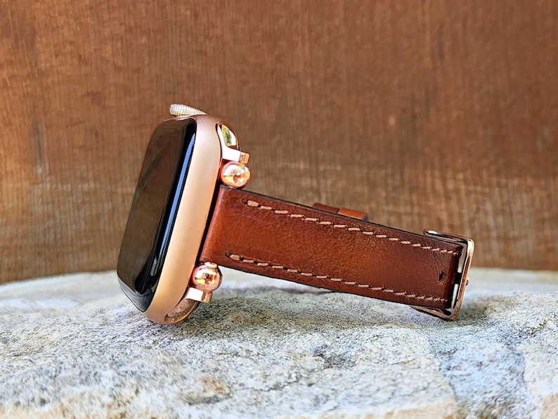 May include: A rose gold smartwatch with a brown leather band. The watch face is black, and the band is stitched with a lighter brown thread. The watch is displayed on a textured, light gray surface.