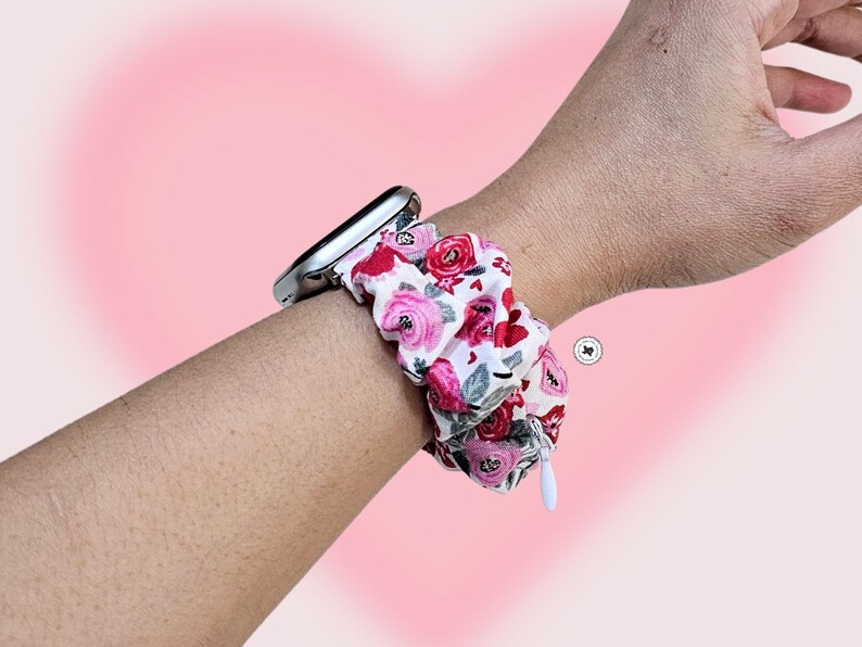 Peut inclure: Bracelet de montre scrunchie floral avec un cadran argent&eacute;. Le scrunchie pr&eacute;sente un fond blanc avec des roses rouges et roses et des feuilles vertes. Une petite fermeture &eacute;clair blanche est visible sur le c&ocirc;t&eacute;.