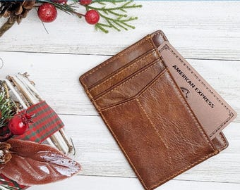 May include: A brown leather cardholder with visible stitching and multiple card slots. An American Express card is partially inserted. The wallet is on a white wooden surface, with holiday decorations in the background.