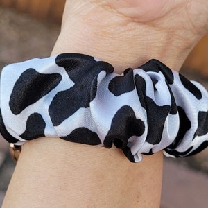 Op de afbeelding: Een stoffen haarelastiek met een herhalend zwart-wit koeienvlekkenpatroon. Het accessoire is gemaakt van een glad, licht glanzend materiaal en is in een cirkelvorm geplooid met een elastische binnenkant om het haar vast te zetten.