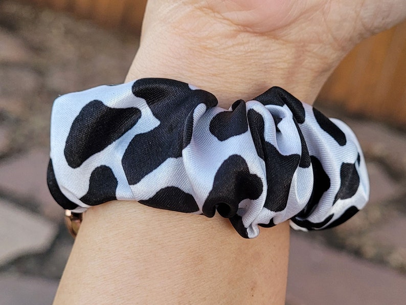 Op de afbeelding: Een stoffen haarelastiek met een herhalend zwart-wit koeienvlekkenpatroon. Het accessoire is gemaakt van een glad, licht glanzend materiaal en is in een cirkelvorm geplooid met een elastische binnenkant om het haar vast te zetten.