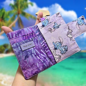 May include: A fabric wallet featuring a purple batik pattern on one side and a light purple fabric with a repeating blue alien character print on the other. The wallet includes multiple card slots and a small tag with the text Roe Allen.