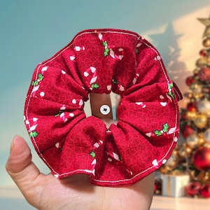 May include: A red fabric hair scrunchie featuring a repeating pattern of white and red candy canes with small green holly leaves. The scrunchie has visible white stitching along the outer edge and a small circular white logo tag in the center.