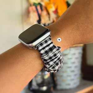 May include: A silver-toned smartwatch featuring a black screen and a fabric scrunchie-style watch band. The band displays a classic black and white gingham checkered pattern with a gathered, elasticized texture that wraps around the wrist.