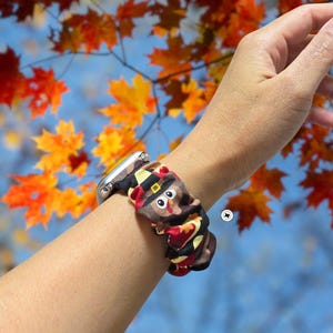 Op de afbeelding: Een zilveren smartwatch met een patroonband met een kalkoenontwerp. De band is bruin, rood en geel, met een cartoon kalkoengezicht. De achtergrond toont herfstbladeren in oranje en gele tinten.