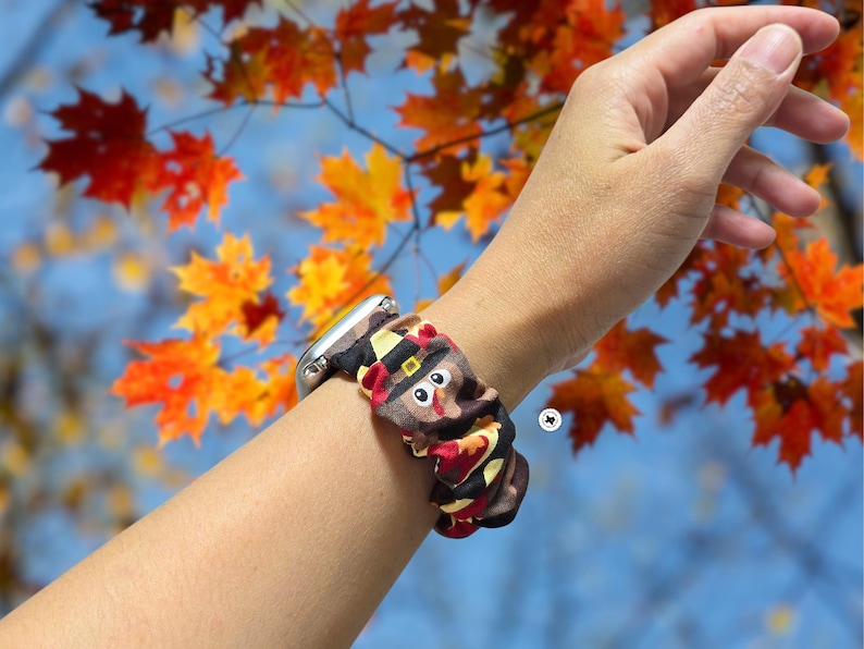 Op de afbeelding: Een zilveren smartwatch met een patroonband met een kalkoenontwerp. De band is bruin, rood en geel, met een cartoon kalkoengezicht. De achtergrond toont herfstbladeren in oranje en gele tinten.
