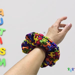 May include: A colorful hair scrunchie with a puzzle piece pattern. The scrunchie is on a person's wrist. The word "AUTISM" is spelled out in colorful letters on a white wall behind the person's arm.