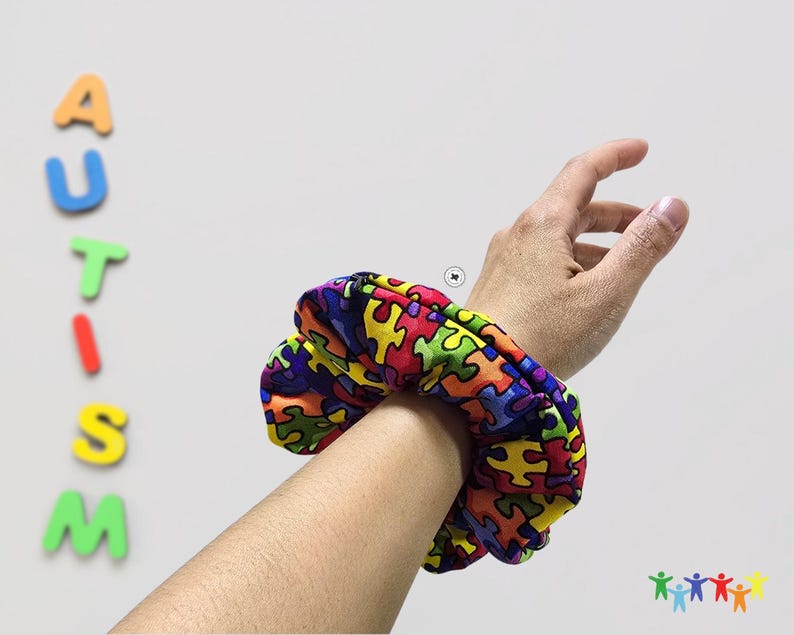 May include: A colorful hair scrunchie with a puzzle piece pattern. The scrunchie is on a person's wrist. The word "AUTISM" is spelled out in colorful letters on a white wall behind the person's arm.