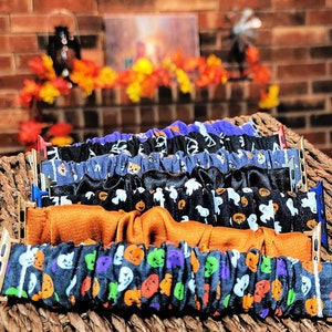 May include: A collection of five fabric scrunchie-style watch bands for smartwatches, featuring various Halloween-themed patterns including pumpkins, ghosts, and solid orange, each attached to metal connectors in gold, silver, and blue.