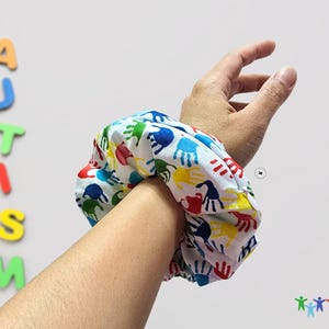 Puede incluir: Un scrunchie blanco con un estampado de huellas de manos de colores. El scrunchie est&aacute; en la mu&ntilde;eca de una persona.