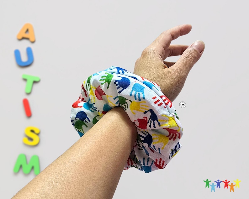 Puede incluir: Un scrunchie blanco con un estampado de huellas de manos de colores. El scrunchie est&aacute; en la mu&ntilde;eca de una persona.