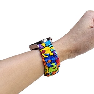 May include: A scrunchie-style fabric watch strap featuring a vibrant, multi-coloured puzzle piece pattern in shades of red, yellow, blue, green, and purple. The elasticated fabric strap is attached to a metallic rose gold smartwatch casing.