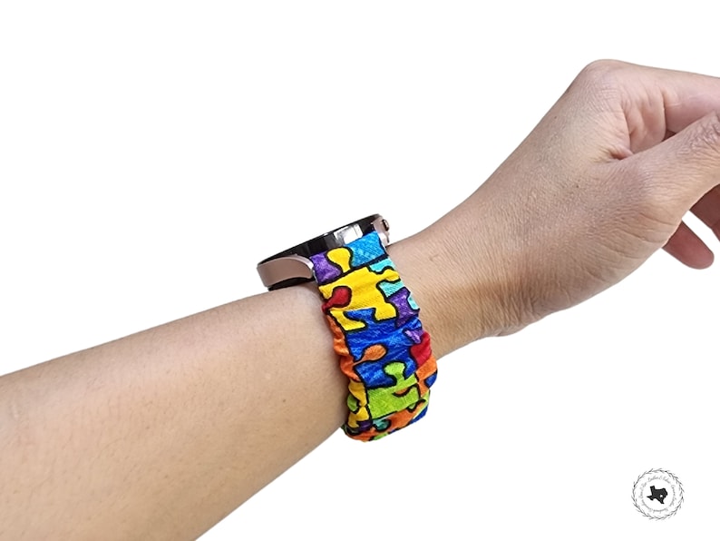 May include: A scrunchie-style fabric watch strap featuring a vibrant, multi-coloured puzzle piece pattern in shades of red, yellow, blue, green, and purple. The elasticated fabric strap is attached to a metallic rose gold smartwatch casing.