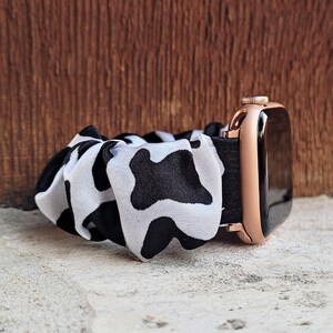 Op de afbeelding: Een horlogeband in scrunchie-stijl met een zwart-wit koeienvlekkenpatroon, bevestigd aan een ros&eacute;gouden smartwatch-behuizing met een zwart scherm. De stof is geplooid en elastisch, wat zorgt voor een zachte, gerimpelde uitstraling om de pols.