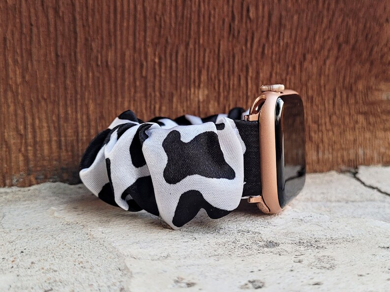 Op de afbeelding: Een horlogeband in scrunchie-stijl met een zwart-wit koeienvlekkenpatroon, bevestigd aan een ros&eacute;gouden smartwatch-behuizing met een zwart scherm. De stof is geplooid en elastisch, wat zorgt voor een zachte, gerimpelde uitstraling om de pols.