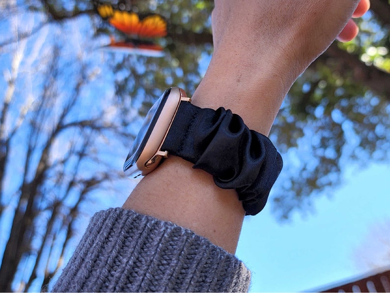 May include: A rose gold smartwatch with a black satin scrunchie strap. The watch is worn on a person's wrist.