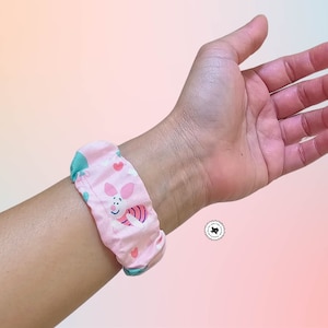 May include: A pink fabric scrunchie with a cartoon pig design, hearts, and white clouds. The scrunchie is worn on a wrist. The scrunchie is made of a soft, patterned fabric. The background is a gradient of pink, blue, and yellow.