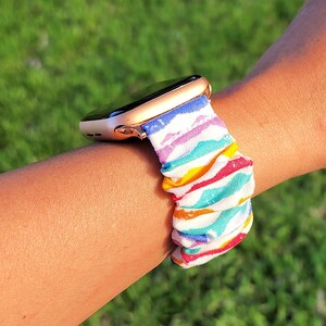 May include: A gold Apple Watch with a rainbow striped fabric band. The band is made of a soft, stretchy material and has a colourful, geometric pattern.