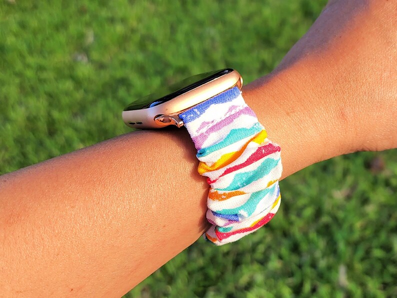 May include: A gold Apple Watch with a rainbow striped fabric band. The band is made of a soft, stretchy material and has a colourful, geometric pattern.