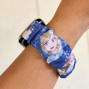 May include: A blue and white patterned watch band featuring a picture of Elsa from Frozen. The band is made of fabric and has a scrunchie-like design.