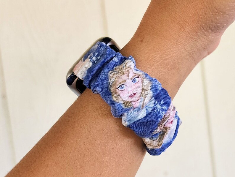 May include: A blue and white patterned watch band featuring a picture of Elsa from Frozen. The band is made of fabric and has a scrunchie-like design.