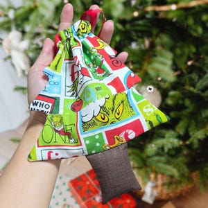 May include: A Christmas tree-shaped ornament made of fabric, featuring a Grinch-themed pattern in red, green, and white. The ornament has a brown rectangular base and a red ribbon for hanging. The design includes the text "WHO" and "ONE".