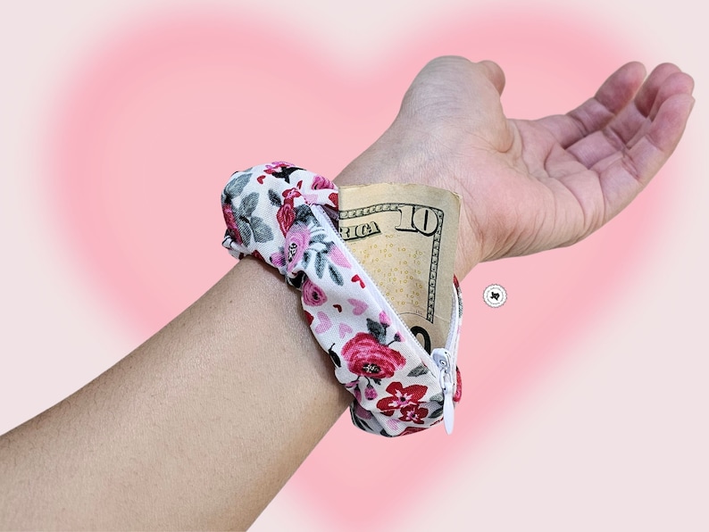 Peut inclure: Un portefeuille de poignet &agrave; imprim&eacute; floral avec une fermeture &eacute;clair blanche, contenant un billet de dix dollars. Le tissu pr&eacute;sente un motif de roses rouges et roses sur fond blanc. Le bracelet est con&ccedil;u pour &ecirc;tre port&eacute; autour du poignet.