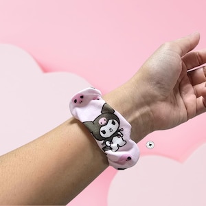 Op de afbeelding: Een lichtroze scrunchie met een herhalend patroon van een stripfiguur met een zwart en bruin hoofd en een wit lichaam. De scrunchie is om een pols gewikkeld. De achtergrond is lichtroze.
