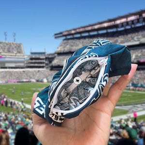 Puede incluir: Una bolsa con cremallera de color verde azulado y blanco con un dise&ntilde;o de logotipo de equipo de f&uacute;tbol. La bolsa est&aacute; abierta, revelando un interior estampado en gris y blanco. El fondo muestra un estadio de f&uacute;tbol.