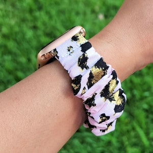 Op de afbeelding: Een horlogeband in scrunchie-stijl met een lichtroze stoffen basis en een abstract patroon van zwarte en metallic gouden vlekken. De band is bevestigd aan een ros&eacute;gouden horlogekast, wat zorgt voor een geplooide en elastische uitstraling.