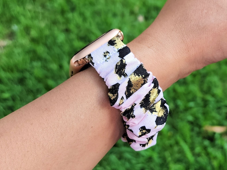 Op de afbeelding: Een horlogeband in scrunchie-stijl met een lichtroze stoffen basis en een abstract patroon van zwarte en metallic gouden vlekken. De band is bevestigd aan een ros&eacute;gouden horlogekast, wat zorgt voor een geplooide en elastische uitstraling.