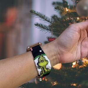 May include: A gold smartwatch with a black and white patterned band featuring a green cartoon character. The band has red and white polka dots. The watch is worn on a person's wrist near a Christmas tree.