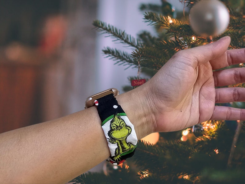 May include: A gold smartwatch with a black and white patterned band featuring a green cartoon character. The band has red and white polka dots. The watch is worn on a person's wrist near a Christmas tree.