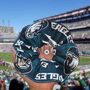 Puede incluir: Una scrunchie verde azulado y blanca con el logotipo y el nombre del equipo de los Philadelphia Eagles. La scrunchie se sostiene frente a un estadio de f&uacute;tbol.